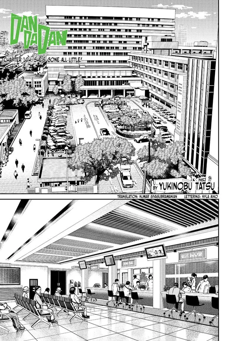 An open-air parking lot in front of a huge hospital building with many cars and people walking.